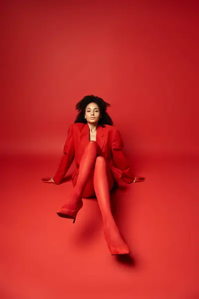 Young woman wearing a red jacket and tights, sitting with legs crossed on a vibrant red floor in a studio setting. — Stock Photo