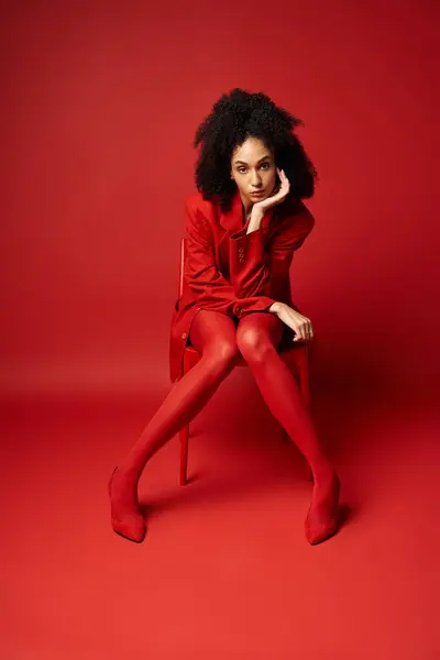 A young woman in a stunning red jacket and tights is gracefully sitting on a chair against a vibrant studio backdrop. — Stock Photo
