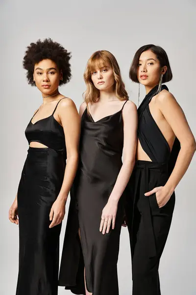 Tre donne di etnie diverse che si riuniscono in abiti neri su uno sfondo grigio, mostrando diversità ed eleganza. — Foto stock