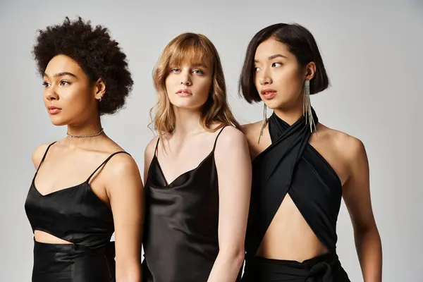 Три жінки різних національностей витончено стоять у чорних сукнях на фоні нейтральної сірої студії. — стокове фото