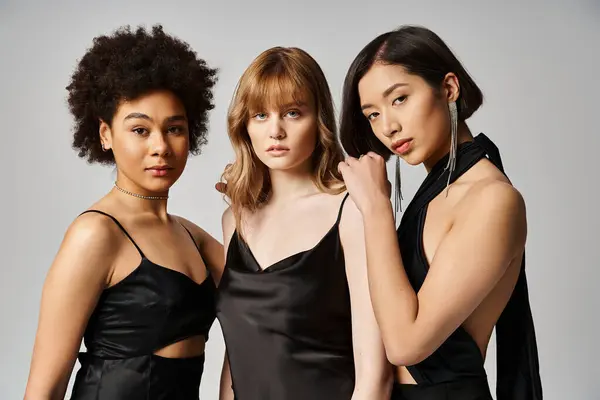 Trois belles femmes d'ethnies différentes dans des robes noires élégantes posant gracieusement sur un fond gris. — Photo de stock