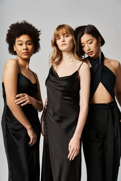 Três mulheres de diferentes etnias vestindo vestidos pretos ficam graciosamente ao lado umas das outras contra um fundo de estúdio cinza. — Fotografia de Stock