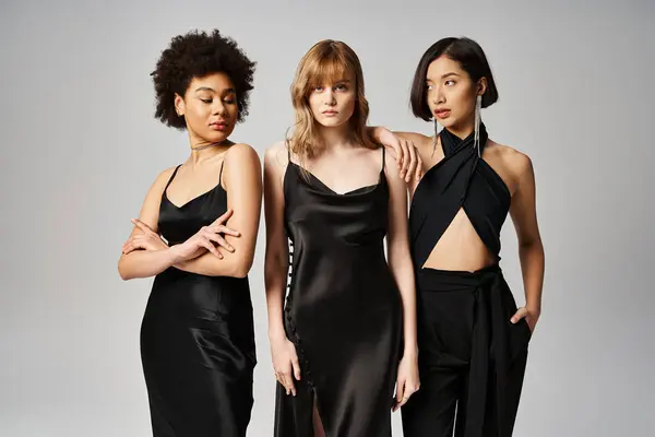 Tres mujeres, cada una con un elegante vestido negro, se paran elegantemente juntas sobre un fondo gris de estudio. - foto de stock