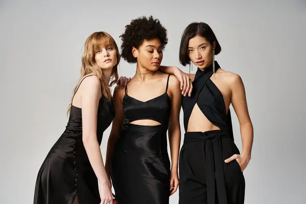 Three women of different ethnicities - Caucasian, Asian, African American - standing in black dresses against a grey background. — Stock Photo