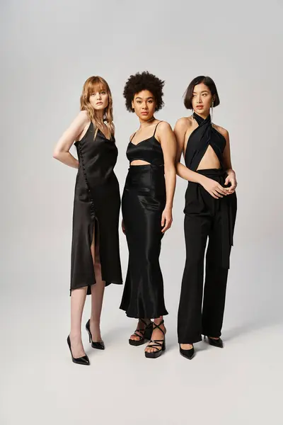 Три красиві жінки різних національностей стоять поруч, одягнені в шикарні чорні вбрання на тлі сірого студійного фону. — стокове фото