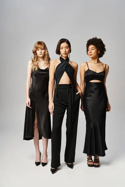 Three women of diverse backgrounds stand next to each other in elegant black dresses on a grey studio background. — Stock Photo