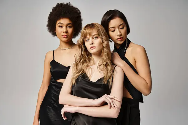Tres elegantes mujeres de diferentes razas se paran elegantemente juntas contra un fondo gris. - foto de stock