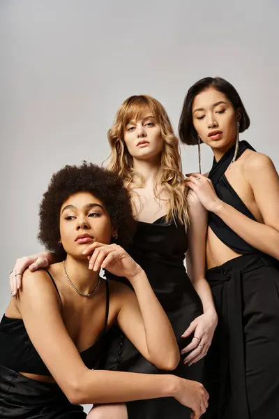 Tres mujeres de diversos orígenes en vestidos negros posan con gracia en un estudio sobre un fondo gris. - foto de stock