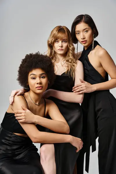 Три жінки в чорних сукнях витончено стоять, демонструючи різноманітність і красу з сірим студійним фоном. — стокове фото