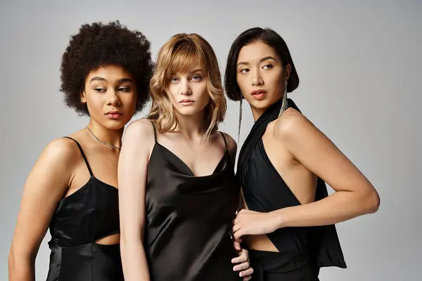 Tre donne vestite di nero posano per una foto sullo sfondo di uno studio grigio, mettendo in mostra bellezza e diversità. — Foto stock