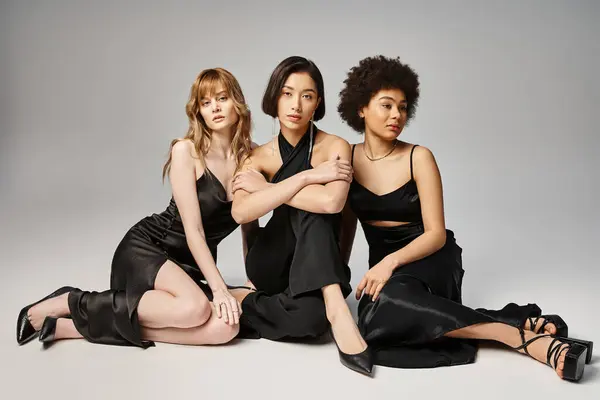 Three women of different ethnicities sit gracefully next to each other in a studio setting against a grey background. — Stock Photo