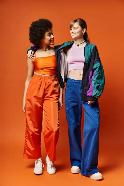 Due diverse giovani donne di discendenza caucasica e afroamericana in piedi insieme sullo sfondo di uno studio arancione. — Foto stock