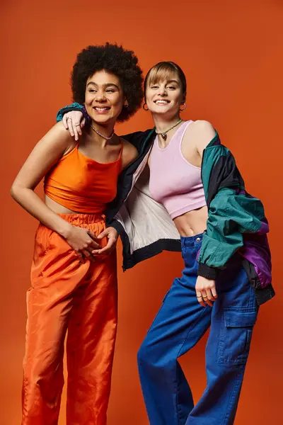 Duas belas mulheres de diferentes etnias estão lado a lado em um estúdio, sorrindo e posando para uma foto contra um fundo laranja. — Fotografia de Stock