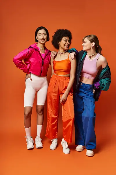 Drei schöne Frauen kaukasischer, asiatischer und afroamerikanischer Abstammung stehen zusammen vor orangefarbenem Studiohintergrund. — Stockfoto