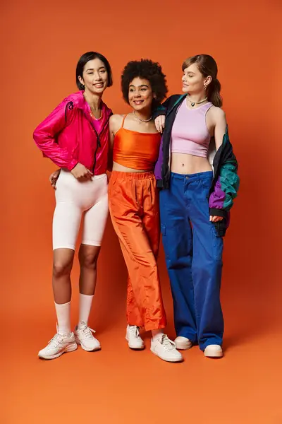 Three women of diverse backgrounds stand united, representing beauty and strength against an orange studio backdrop. — Stock Photo