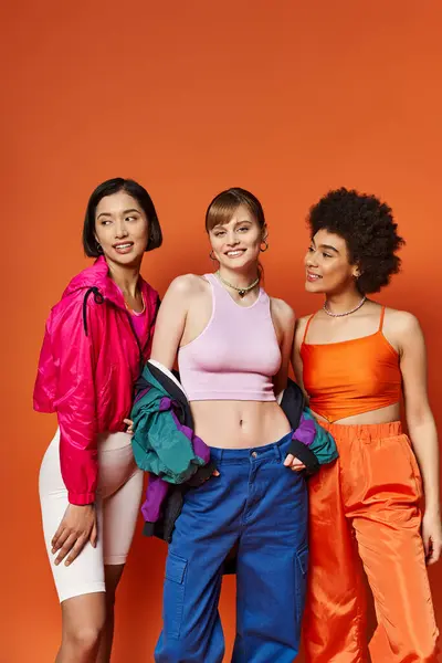A caucasian, asian, and african american woman stand together in a studio against an orange background, showcasing beauty in diversity. — Stock Photo