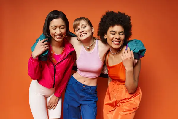 Three diverse women of Caucasian, Asian, and African American descent standing happily together against an orange studio background. — Stock Photo