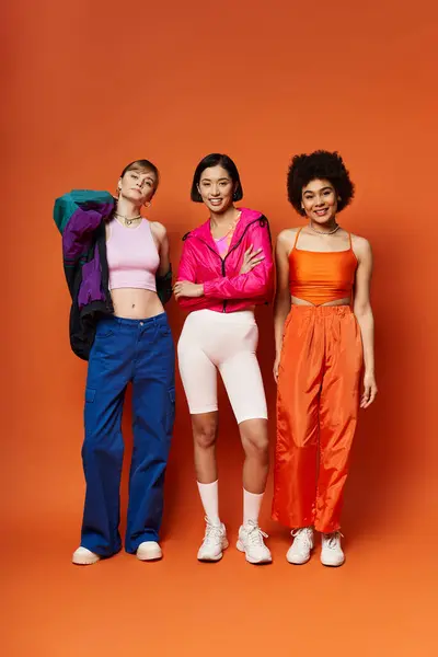 A Caucasian, Asian, and African American woman standing together in front of an orange studio background. — Stock Photo