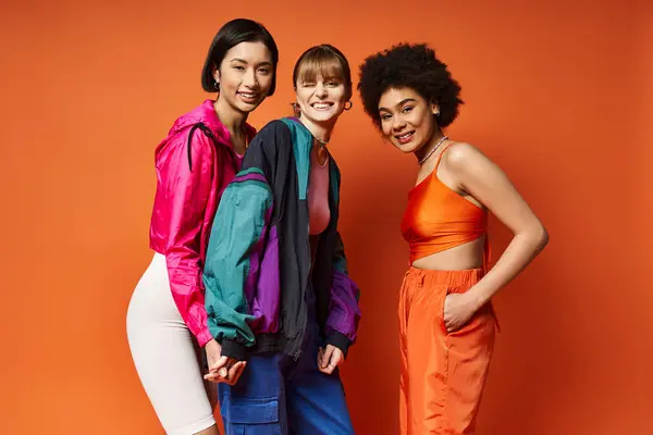 Trois femmes d'ethnies différentes se tenant ensemble dans un studio sur fond orange, rayonnant de beauté et d'unité. — Photo de stock