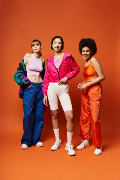 Три красиві жінки в студії, що представляють різноманітність: кавказька, азіатська та афроамериканська, стоять разом на помаранчевому тлі. — стокове фото