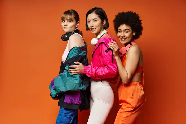 Três mulheres de diferentes etnias e estilos de pé juntos em um estúdio com um fundo laranja vibrante. — Fotografia de Stock