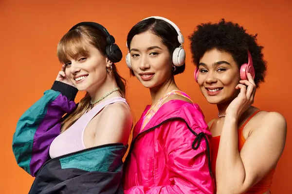 Drei unterschiedliche Frauen mit Kopfhörern posieren im Studio vor orangefarbenem Hintergrund und verkörpern Schönheit und Einheit. — Stockfoto