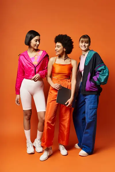 Tres hermosas mujeres de diferentes etnias de pie unidas frente a un fondo naranja. - foto de stock