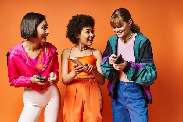 Drei unterschiedliche junge Frauen lachen und starren auf ihre Handys vor einer leuchtend orangen Studiokulisse. — Stockfoto