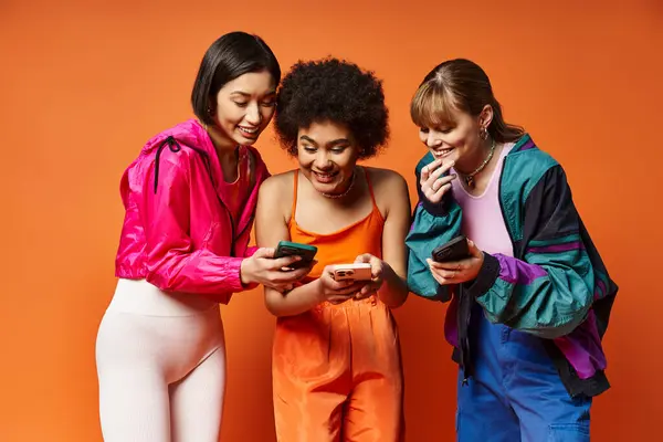 Tres niñas diversas, incluyendo caucásicas, asiáticas y afroamericanas, acurrucadas juntas mirando un teléfono celular con un fondo naranja. - foto de stock