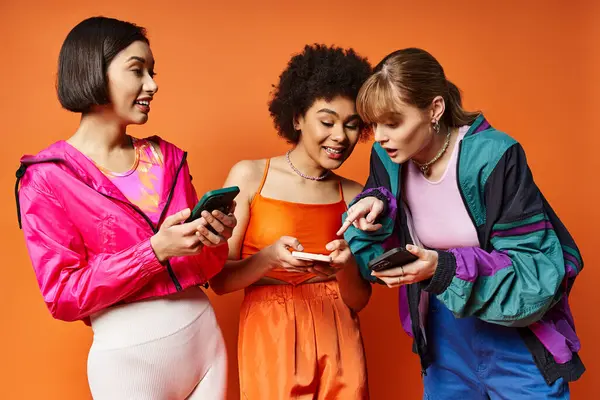 Tre donne diverse con etnie diverse in piedi l'una accanto all'altra, assorbite nei loro telefoni cellulari su uno sfondo arancione. — Foto stock