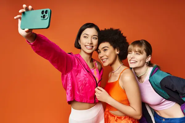 Three women, representing different cultures, enjoying a playful moment as they take a selfie with a cell phone against an orange background. — Stock Photo
