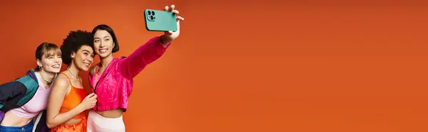 Femmes multiculturelles prenant un selfie sur un téléphone portable sur un fond de studio orange dans un moment joyeux. — Photo de stock