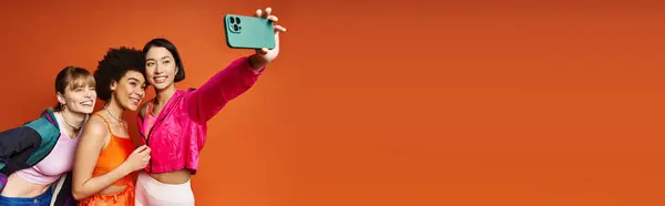 Multicultural women taking a selfie on a cell phone against an orange studio background in a joyful moment. — Stock Photo