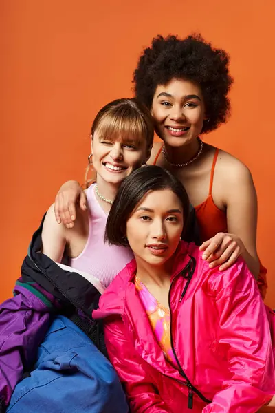Tres mujeres jóvenes de diversos orígenes posando juntas sobre un telón de fondo naranja. - foto de stock