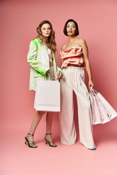 Two beautiful women of different ethnicities standing with shopping bags against a pink background. — Stock Photo