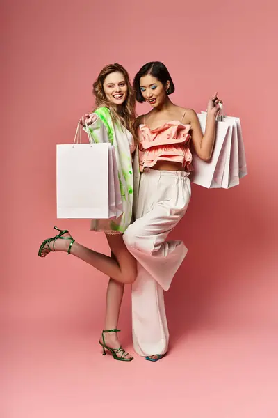 Dos hermosas mujeres jóvenes de diversos orígenes se unen sosteniendo bolsas de compras contra un fondo de estudio rosa. - foto de stock