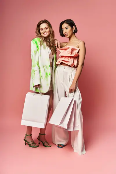 Two multicultural women standing side by side, holding shopping bags on a vibrant pink background. — Stock Photo