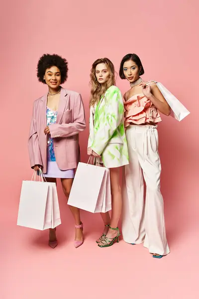 Tre donne di sfondi diversi in piedi insieme, tenendo borse della spesa vivaci su uno sfondo rosa. — Foto stock