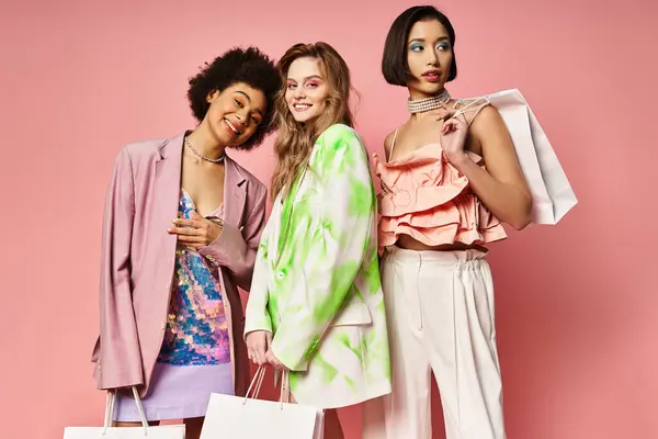 Three women from diverse backgrounds stand together, holding colorful shopping bags in front of a pink studio background. — Stock Photo