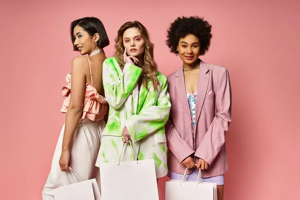 Tres mujeres de diferentes etnias sosteniendo bolsas de compras contra un fondo de estudio rosa. - foto de stock