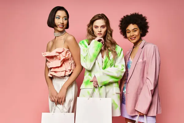 Three diverse women, Caucasian, Asian, and African American, standing together holding shopping bags against a pink background. — Stock Photo