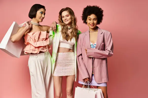 Tres mujeres de diversos orígenes están de pie lado a lado, sosteniendo bolsas de compras contra un vibrante fondo de estudio rosa. - foto de stock