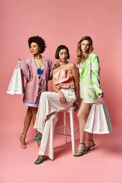 Три разных женщины сидят на стуле, держа сумки с покупками на фоне розовой студии. — стоковое фото