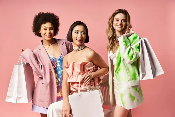 Tres mujeres de diversos orígenes posan elegantemente con bolsas de compras sobre un fondo de estudio rosa. - foto de stock