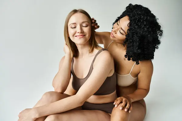 Zwei unterschiedliche Frauen in kuscheliger Pastellunterwäsche sitzen zusammen auf weißem Hintergrund. — Stockfoto
