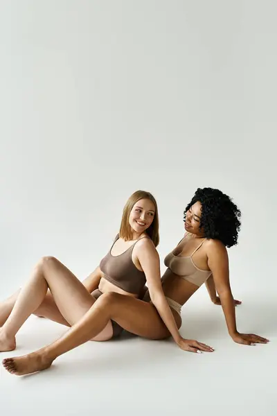 Dos hermosas mujeres diversas se sientan de cerca en el suelo en ropa interior de pastel acogedor. - foto de stock