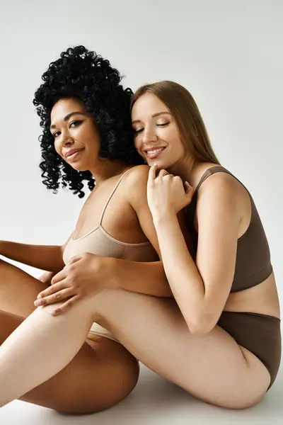 Deux belles femmes diversifiées en sous-vêtements pastel confortables assis les uns à côté des autres sur le sol. — Photo de stock