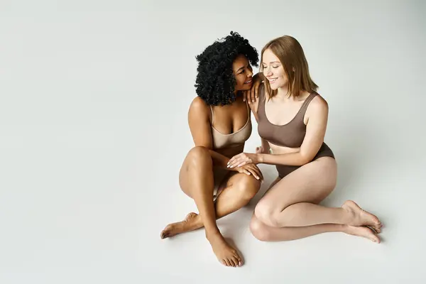 Zwei schöne unterschiedliche Frauen in kuscheliger Pastellunterwäsche, die nebeneinander auf weißem Hintergrund sitzen. — Stockfoto