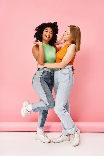 Two women in casual attire embrace, smiling against a pink backdrop. — Stock Photo