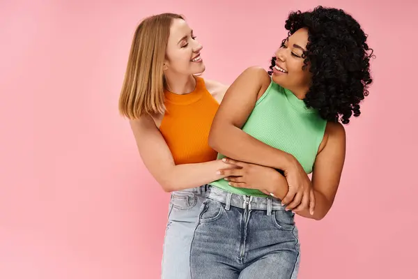 Two attractive diverse women in casual attire hugging in front of a pink background. — Stock Photo